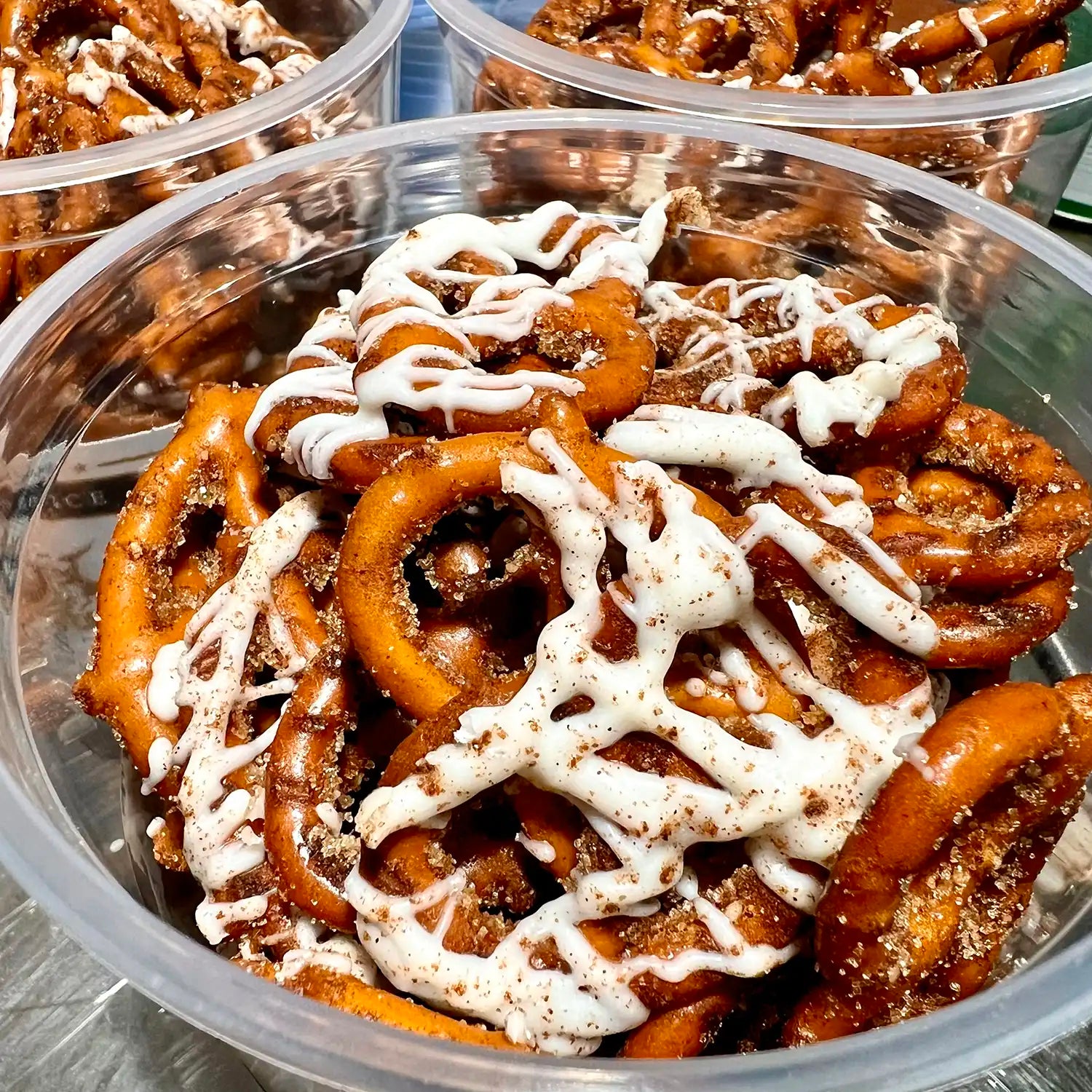 Pretzels with white drizzle in clear plastic containers on a metal surface.